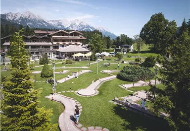 A spacious garden landscape with a mini-golf course and various trees. In the background, mountains and a stylish building can be seen.