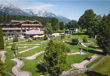 A beautiful mini-golf course with well-maintained lanes and plenty of greenery. In the background, mountains and a hotel can be seen.