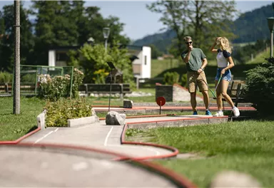 A mini-golf course with green grass and red markings. Two people are strolling leisurely along the playing lanes.