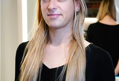 Eine Person mit langen Haaren steht vor einem Spiegel. Sie trägt ein schwarzes Shirt und hat ein freundliches Lächeln.