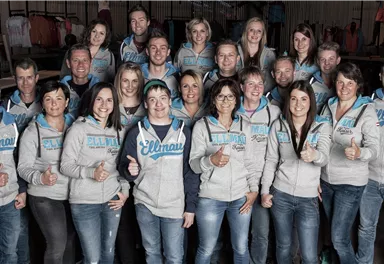 Eine Gruppe von Menschen steht lächelnd zusammen, alle in ähnlichen grauen Sweatshirts. Sie zeigen Daumen hoch und scheinen freundlich und einheitlich.