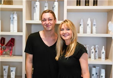 Zwei Personen stehen lächelnd in einem modernen Geschäft. Im Hintergrund sind verschiedene Haarpflegeprodukte sichtbar.