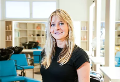 Eine Friseurin steht in einem modernen Salon. Hinter ihr sind frisurentechnische Produkte und Stühle sichtbar.