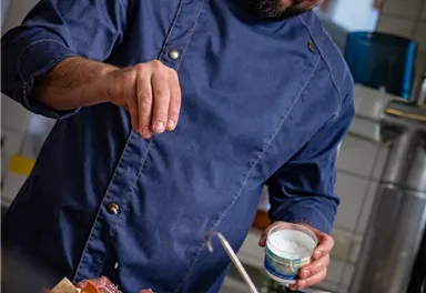 A chef in a modern kitchen seasons fresh vegetables and meat. He is wearing a blue chef's jacket and smiles while preparing the food.