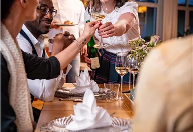 A cheerful dinner scene with guests sitting at a table. A waitress is serving a drink and smiling.