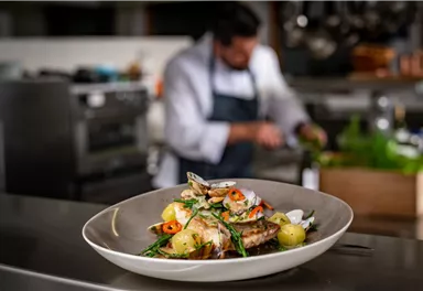 A delicious dish with fish, potatoes, and fresh herbs is on a table. In the background, a chef is working in the kitchen.
