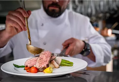 A chef arranges a beautiful dish with pink roasted meat and fresh vegetables. The focus is on the presentation of the food on a white plate.