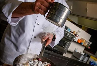 A chef sprinkles powdered sugar over a fresh fruit bowl. In the background, cooking utensils and a modern kitchen can be seen.