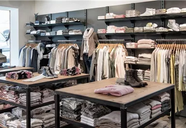 Ein moderner Bekleidungsladen mit Regalen voller Kleidung. Auf den Tischen liegen gefaltete Textilien und Schuhe stehen bereit.
