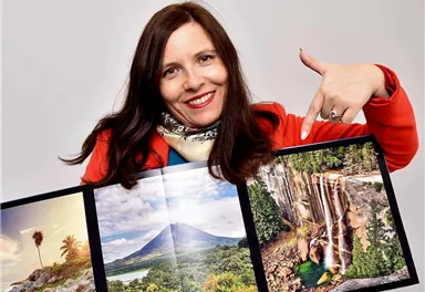 Eine lächelnde Frau hält ein Fotobuch mit Bildern von Natur und Wasserfällen. Sie zeigt auf die beeindruckenden Landschaften im Buch.