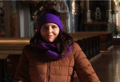 Eine Frau mit langen Haaren trägt einen lila Hut und einen braunen Mantel. Sie steht in einem dekorativen Innenraum mit Holzdetails.