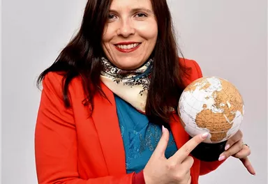 Eine lächelnde Frau in einem roten Blazer hält einen Globus in der Hand. Sie zeigt auf den Globus und trägt einen Schal um den Hals.