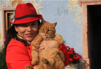 Eine Frau mit einem roten Hut hält eine orangefarbene Katze in ihren Armen. Im Hintergrund sind blühende Pflanzen und eine farbige Wand zu sehen.