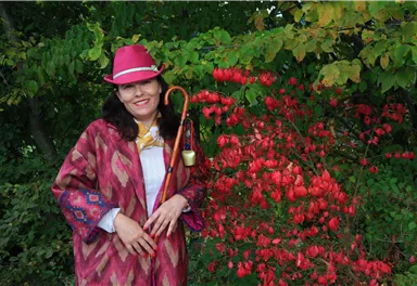 Eine lächelnde Frau in einem bunten Outfit steht neben einem blühenden Strauch mit roten Früchten. Sie trägt einen Hut und hält einen Gehstock in der Hand.