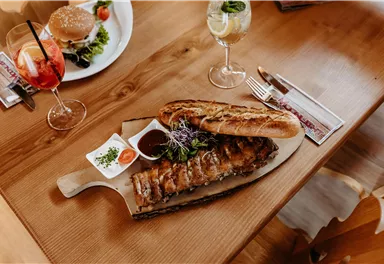 A delicious dish of grilled meat on a wooden board, served with bread and dip. In the background, cocktails and a salad can be seen.