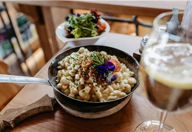 A plate of creamy pasta, garnished with herbs and edible flowers. In the background, there is a small salad and a glass of drink.