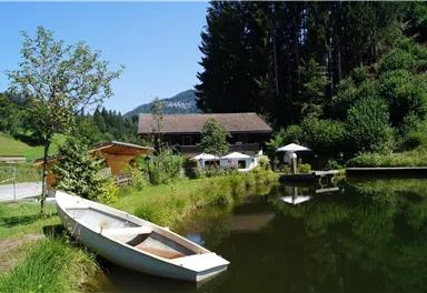 A calm pond with a boat and surrounding green meadows. In the background, a wooden house and some sun umbrellas can be seen.