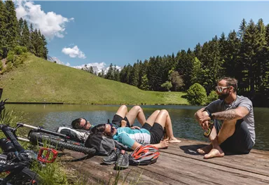Three people are relaxing by the shore of a calm lake. In the background, green meadows and dense forests can be seen.