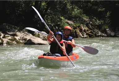 Two people are paddling in a red kayak on a wild river. They are wearing helmets and seem to be having a lot of fun.
