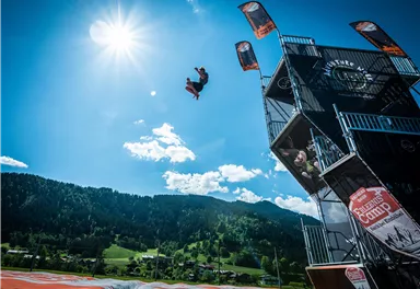 An athlete jumps from a high platform into the sky. In the background, green hills and a clear blue sky can be seen.