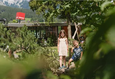 Two children are standing in a garden with trees and plants. In the background, a modern building view can be seen.