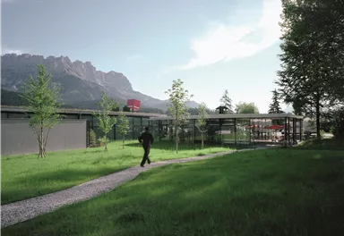 A modern building in a green landscape with mountains in the background. A man is walking along a path that leads through the grass.