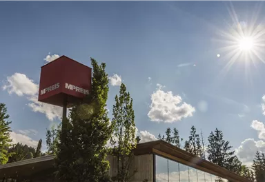 A modern building with a large window front and a red sign. Bright sunshine and blue skies with some clouds in the background.