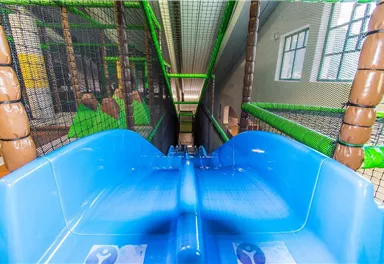 A blue slide in an indoor play area with nets and colorful walls. The view is directed downward from the slide's perspective.