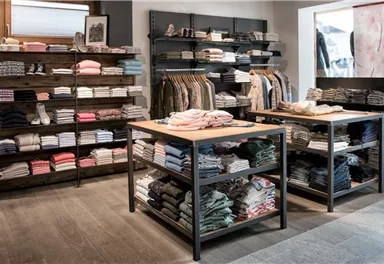 Ein moderner Bekleidungsladen mit ordentlich gestapelten Kleidungsstücken auf Regalen und Tischen. Die Farbpalette umfasst sanfte Töne und schafft eine einladende Atmosphäre.