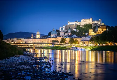 A picturesque view of the city of Salzburg at night. The lights reflect in the river, while the fortress shines in the background.