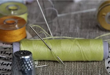 Eine Nähspule mit grünem Garn und einer Nadel liegt auf einem Tisch. Im Hintergrund sind weitere Nähspulen in verschiedenen Farben zu sehen.