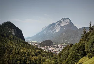 Eine malerische Berglandschaft mit grünen Wäldern und schroffen Gipfeln. Im Tal ist eine kleine Stadt sichtbar.