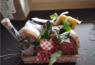 A pretty gift basket with various types of cheese, a bottle of vinegar, and fresh herbs. The basket is decorated with straw and creates an inviting atmosphere.