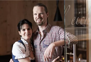 Ein lächelndes Paar steht vor einer Glaswand. Der Mann trägt ein kariertes Hemd, die Frau ein traditionelles Kleid.