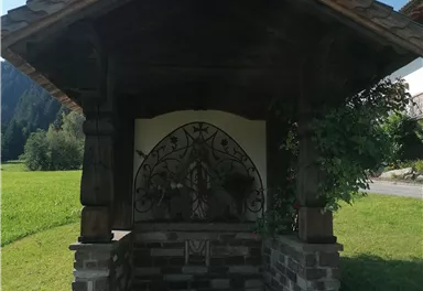 A small chapel with a slanted wooden roof and a cross. It stands in a green meadow surrounded by trees.