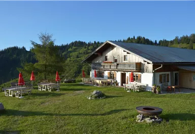 A cozy mountain cabin surrounded by green meadows and trees. Red sun umbrellas and tables invite you to linger.