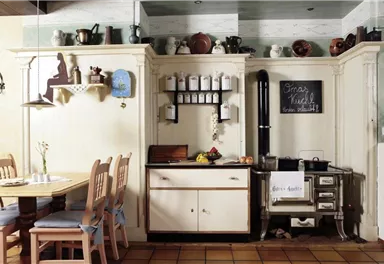 Eine gemütliche Küche mit einem Holztisch und Stühlen. An der Wand befinden sich Regale mit Töpfen und Kräutern sowie ein Herd.