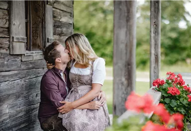 A couple sits by a wooden building and kisses each other. In the foreground, red geraniums are blooming.
