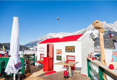 A cozy wooden house with a red roof in a snow-covered mountain landscape. The sky is clear blue and various lights adorn the area.