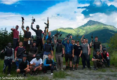 Eine Gruppe von Menschen posiert im Freien mit Gewehren, umgeben von einer hügeligen Landschaft. Der Himmel ist blau mit einigen Wolken.