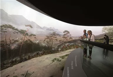 A modern observation platform with visitors offers a view of a picturesque landscape. In the background, mountains and trees can be seen, creating a calm and natural atmosphere.