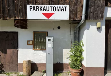 A parking meter next to a building with a sign above it. In the background, a wall and a pot with plants are visible.