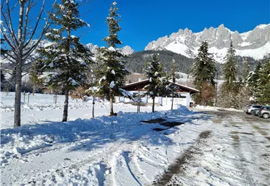 Eine verschneite Landschaft mit hohen Bäumen und einem Chalet. Im Hintergrund ragen majestätische Berge unter einem klaren blauen Himmel auf.