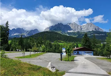 A picturesque parking lot with a view of majestic mountains and a green landscape. The sky is partly cloudy and blue.