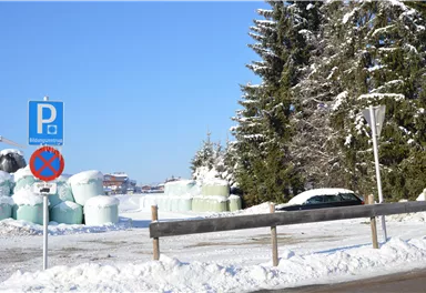 A snowy landscape with a parking sign and a no-entry sign. In the background, large green bales and snow-covered trees can be seen.