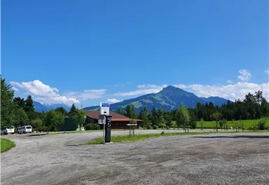 A rural scene with a parking lot and a building in the foreground. In the background, green hills and a large mountain can be seen under a blue sky.