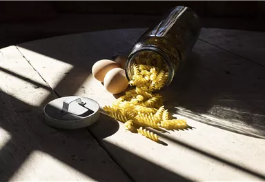 Ein Glas mit spiraligen Nudeln, das auf einem Tisch umgekippt ist. Daneben liegen zwei Eier und eine Dose.