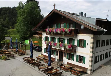 Ein charmantes, traditionelles Gasthaus mit Blumenkästen. Im Außenbereich befinden sich Tische und Sonnenschirme, umgeben von grüner Natur.