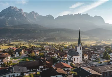 A picturesque Alpine landscape with a charming village view. In the foreground stands a church with a pointed tower, surrounded by mountains and meadows.
