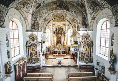 Eine prachtvolle Kirche mit historischen Wandmalereien und einer beeindruckenden Altaranlage. Die Bänke sind bereit für die Besucher, und das Gesamtbild strahlt eine ruhige Atmosphäre aus.